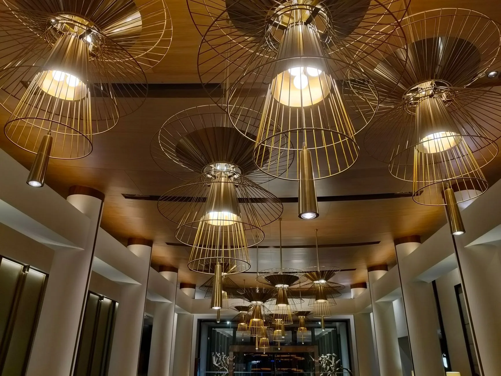 Golden wireframe pendant luminaires layered across the ceiling of a modern hotel lobby, an example of hotel commercial lighting specification